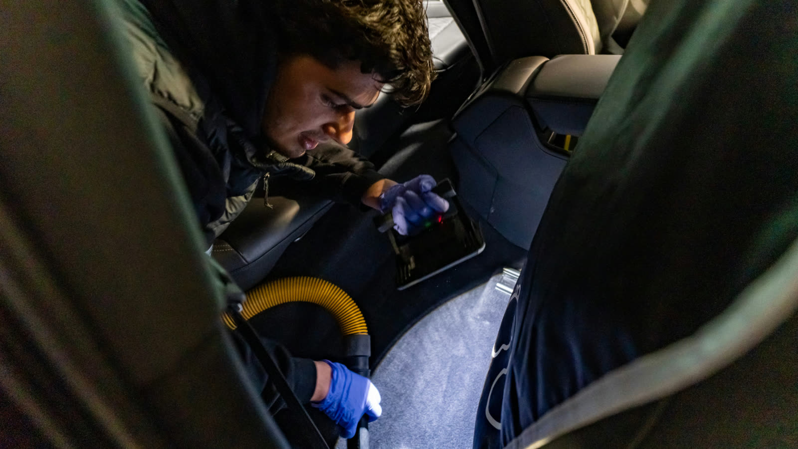 ALPHASHINE detailer vacuuming the center console area of a luxury SUV interior using a yellow hose, wearing gloves and focused on tight spaces.