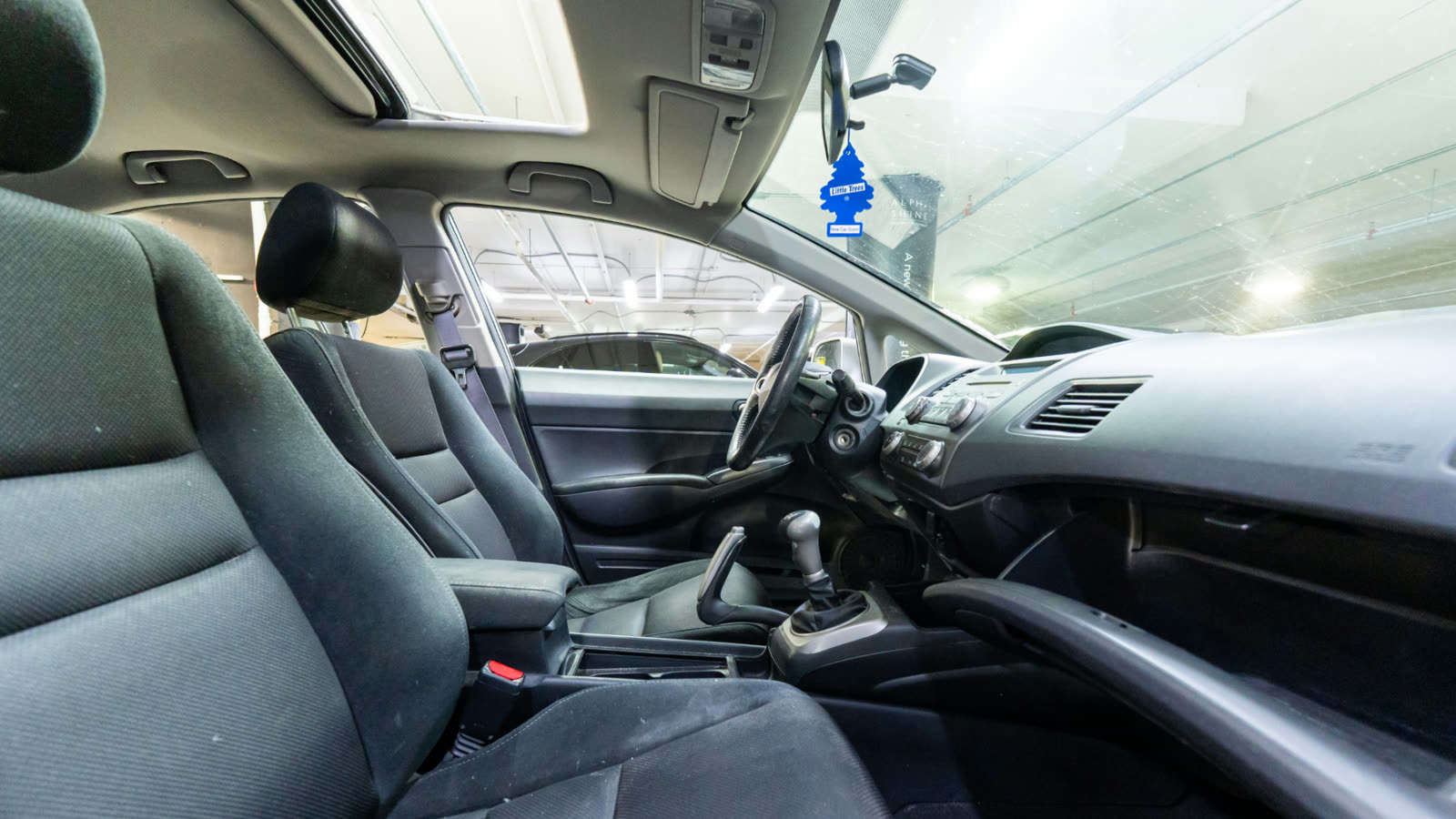 Clean car interior with fabric seats, center console, and manual gear shift, parked inside an underground garage with bright overhead lighting.