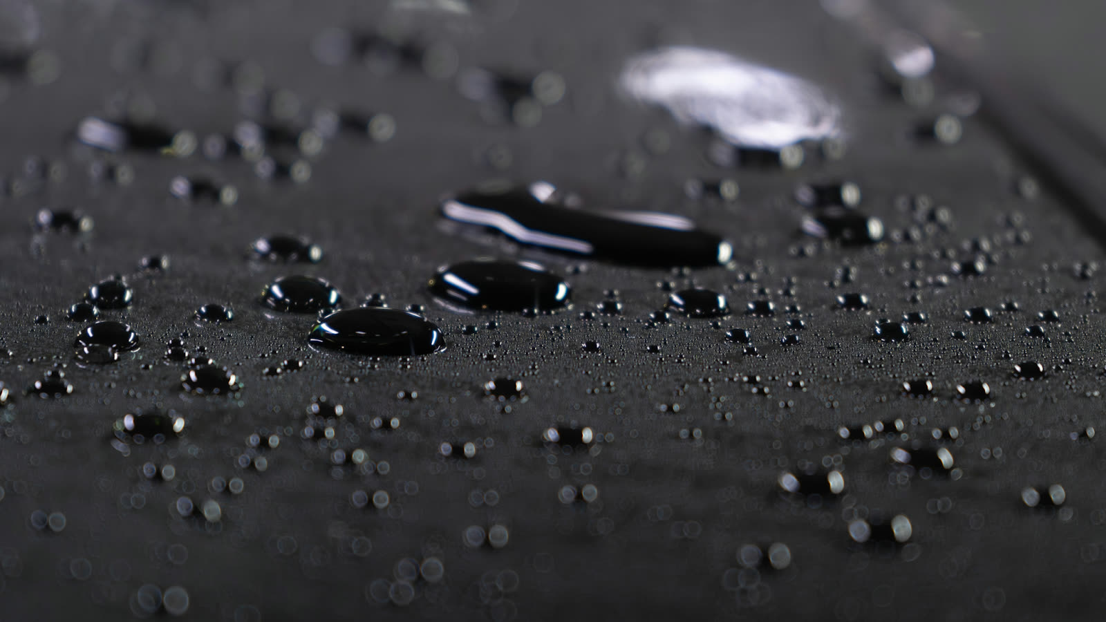 Close up of water droplets beading on a dark car surface, highlighting a smooth, glossy finish with shallow depth of field.
