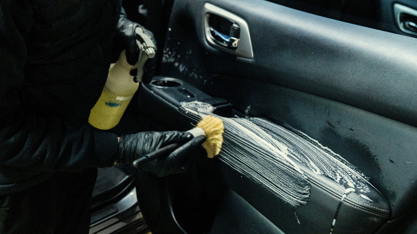 Detailer wearing black gloves sprays cleaner and uses a soft brush to scrub soap onto the interior door panel of a car.