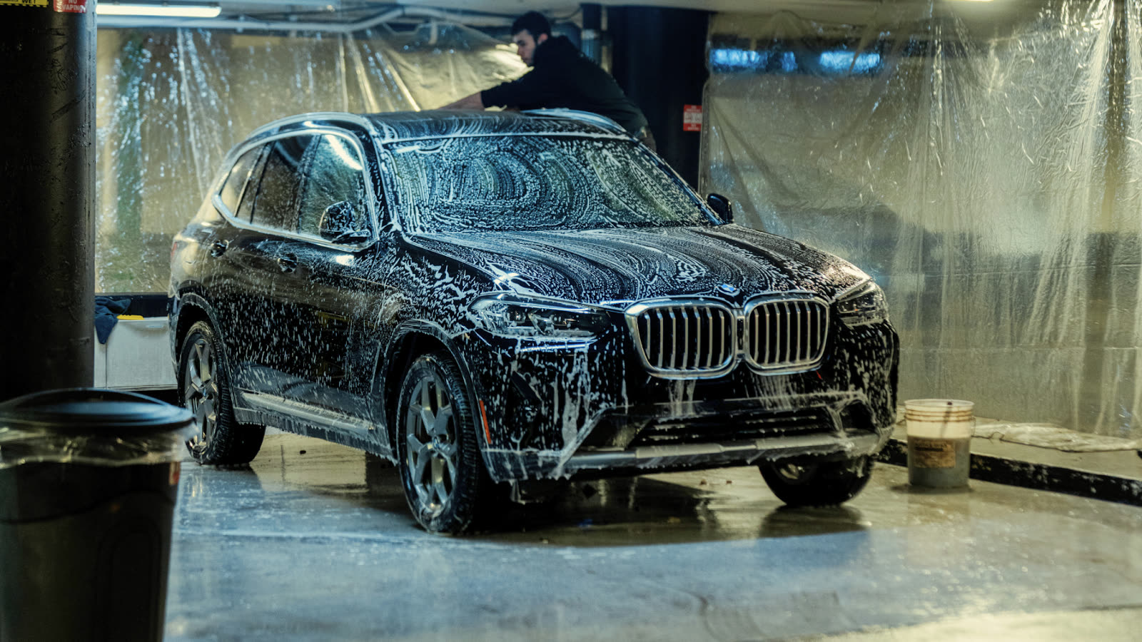 Black SUV covered in white foam during a hand wash inside a garage, with a worker cleaning the roof under bright overhead lights.