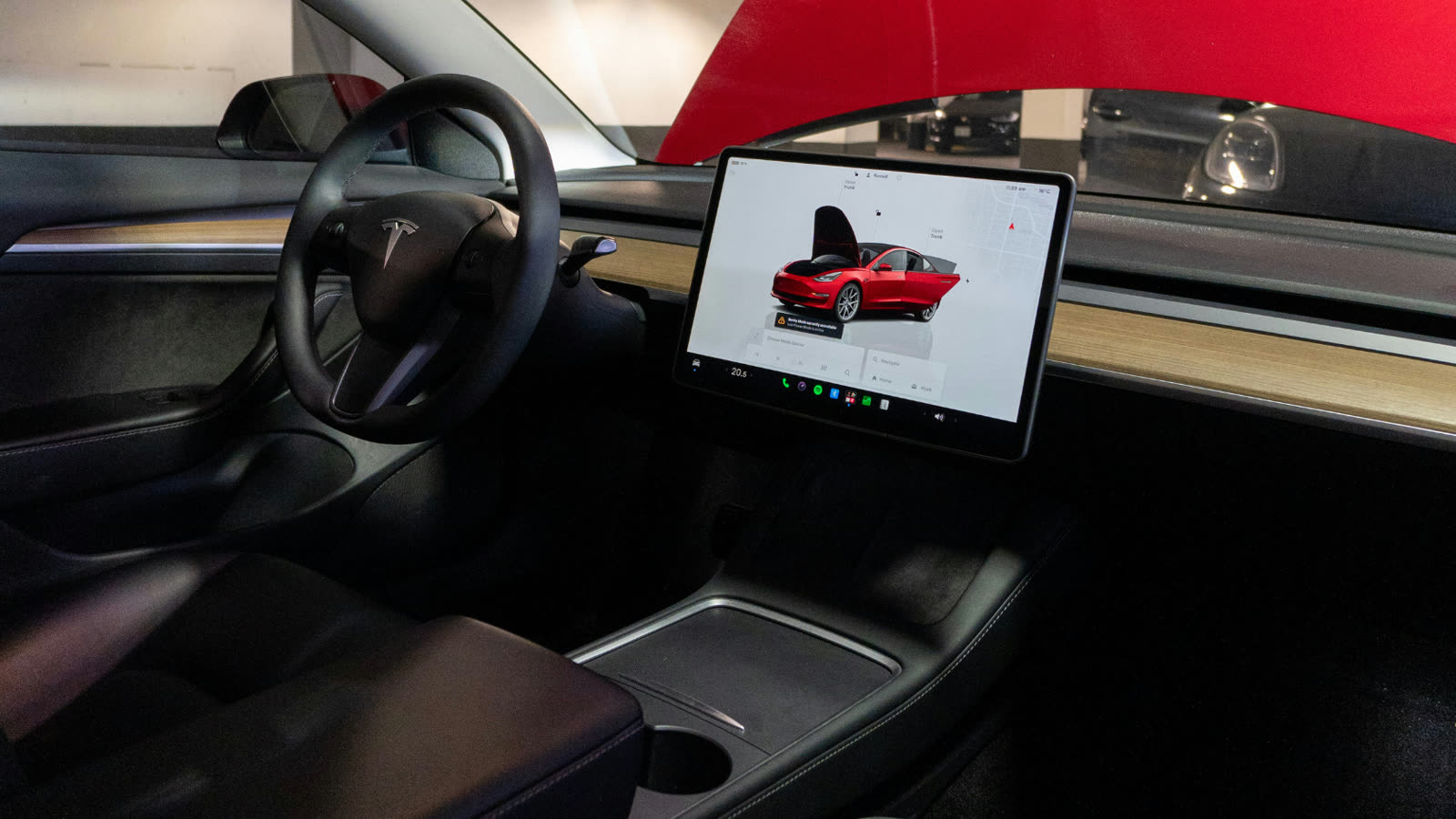 he interior of a Tesla Model 3 showing the minimalist dashboard with a large touchscreen display in the center. The screen shows a 3D graphic of a red Tesla with the front trunk open alongside a navigation map. The flat bottom steering wheel with the Tesla logo is visible on the left and the center console with a wireless charging pad and cup holder is in the foreground.